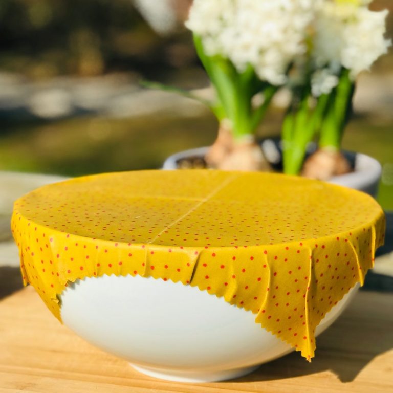 Eine weiße Schüssel mit einem gelben, gepunkteten Bienenwachstuch abgedeckt, im Hintergrund Pflanzen.