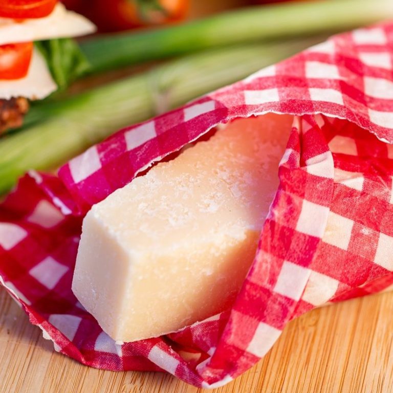 Stück Hartkäse in kariertem roten Bienenwachstuch, umgeben von frischem Gemüse und Kräutern.