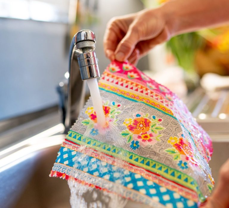 Eine Hand hält ein buntes Bienenwachstuch unter fließendem Wasser am Wasserhahn. So werden Bienenwachstücher gereinigt.