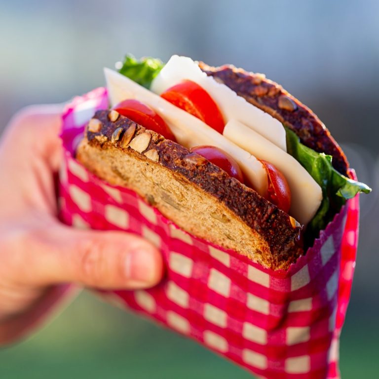 Ein handgehaltenes Sandwich mit Salat, Tomaten und Käse in einem karierten Bienenwachstuch.